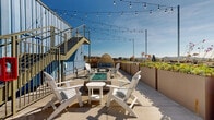 Located at 521 W Colorado St in Glendale, California, this rooftop terrace offers a well-appointed outdoor retreat with panoramic views of the surrounding Glendale cityscape and the San Gabriel Mountains beyond. The terrace is finished with smooth concrete pavers and bordered by a row of large raised planters filled with lush succulents, flowering plants, and greenery, adding a natural, garden-like character to the elevated setting.

At the heart of the space is a built-in fire pit with a glass-bead bed, surrounded by white Adirondack chairs and a side table, creating an inviting lounge area ideal for gathering. A wicker pod daybed provides an additional seating option, offering a cozy nook with cushioned comfort and open-air views. Overhead, strands of globe string lights are strung across the terrace, lending a warm ambiance suited for evening use.

The terrace is set against a bold blue corrugated metal façade, with a metal staircase providing access to an upper level. The industrial architectural elements contrast effectively with the relaxed, resort-style furnishings. The open layout provides generous space for both lounging and social gathering, making this rooftop a functional and visually striking amenity with commanding views under the Southern California sky.