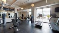Located at 29 St. Helens Avenue in Tacoma, Washington, this well-appointed fitness center offers a comprehensive workout environment designed to accommodate a wide range of exercise needs. The space is organized into distinct functional zones, each catering to different training disciplines.

The cardio area is generously equipped with treadmills, stationary bikes, a spin bike, ellipticals, and a recumbent bike, all positioned to take advantage of the large floor-to-ceiling windows and sliding glass doors that open to an outdoor terrace. These expansive windows flood the space with natural light and frame impressive views of the surrounding landscape, including what appears to be a waterfront panorama.

The strength training section features a robust selection of selectorized machines, a cable system, a full dumbbell rack, and a leg press machine, all set on durable rubber flooring. A mirrored wall in this zone enhances the sense of depth and assists with proper form.

A separate functional training area, enclosed by sleek black-framed glass partition doors, provides a dedicated space for dynamic exercises. It includes kettlebells in various weights, stability balls, and a hardwood floor suitable for stretching or bodyweight movements. The industrial-style exposed ceilings with visible ductwork and pendant globe lighting give the space a modern, urban aesthetic throughout.