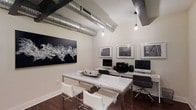 This single-floor workspace showcases a striking blend of industrial and contemporary design elements. Exposed silver ventilation ducts run prominently across the ceiling, complementing the raw, loft-inspired aesthetic. Pendant bulb lighting hangs at varying heights, providing warm, focused illumination throughout the space.

The room features a central white high-gloss table with sleek chrome-legged chairs, serving as a collaborative or multi-use work surface. Along one wall, a dedicated computer workstation area hosts multiple desktop monitors and office equipment, supported by a continuous white desk surface with ergonomic task chairs. The dark hardwood flooring provides a rich contrast to the predominantly white furnishings and walls.

Artistic touches elevate the space considerably — a large-scale black-and-white abstract canvas anchors one wall, while a curated series of framed black-and-white photographic prints lines the adjacent wall above the workstations. The neutral, monochromatic color scheme throughout creates a cohesive, professional atmosphere. Overall, the space balances functional utility with thoughtful design, making it well-suited for focused work or collaborative tasks.