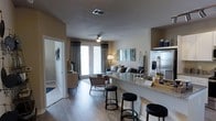 This single-story residence showcases an open-concept layout that seamlessly connects the kitchen and living areas. The kitchen features a prominent granite-topped breakfast bar with seating for multiple bar stools, providing both a functional prep space and a casual dining area. A stainless steel refrigerator with a water and ice dispenser anchors the kitchen, complemented by warm cabinetry above for additional storage.

The living area extends naturally from the kitchen, finished with wood-style plank flooring that runs throughout the space, adding warmth and cohesion. A ceiling fan with an integrated light fixture serves the living room, which is furnished with a sofa, accent chairs with nail-head detailing, and layered rugs. Decorative wall art and framed panels contribute to a contemporary aesthetic.

A set of French doors at the far end of the living room draws in natural light and provides direct access to an outdoor patio or balcony, framed by floor-length curtains in a neutral tone. A hallway visible on the left suggests additional rooms beyond the main living space, completing this well-appointed, efficiently designed residence.