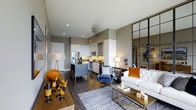 This thoughtfully designed single-floor residence at 1805 offers a seamless open-concept layout that effortlessly connects the kitchen, dining, and living areas. The kitchen is outfitted with stainless steel appliances, including a French door refrigerator and microwave, complemented by sleek white cabinetry and a granite or stone-topped island with bar seating — ideal for casual dining and entertaining. Rich, dark hardwood flooring flows throughout the space, grounding the contemporary aesthetic and providing a warm contrast to the neutral gray walls and white ceilings. Recessed lighting illuminates the space with a clean, modern feel. The living area is generously proportioned, accommodating a full sofa arrangement and accented by a striking floor-to-ceiling industrial-style steel and glass partition that adds an architectural focal point and a touch of urban sophistication. Natural light filters in to enhance the open, airy atmosphere. The overall ambiance is modern and curated, blending warm tones — amber, navy, and gold — with clean lines and high-quality finishes, resulting in a space that feels both stylish and functional.
