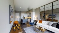 This thoughtfully designed single-floor residence at 1805 offers a seamless open-concept layout that effortlessly connects the kitchen, dining, and living areas. The kitchen is outfitted with stainless steel appliances, including a French door refrigerator and microwave, complemented by sleek white cabinetry and a granite or stone-topped island with bar seating — ideal for casual dining and entertaining. Rich, dark hardwood flooring flows throughout the space, grounding the contemporary aesthetic and providing a warm contrast to the neutral gray walls and white ceilings. Recessed lighting illuminates the space with a clean, modern feel. The living area is generously proportioned, accommodating a full sofa arrangement and accented by a striking floor-to-ceiling industrial-style steel and glass partition that adds an architectural focal point and a touch of urban sophistication. Natural light filters in to enhance the open, airy atmosphere. The overall ambiance is modern and curated, blending warm tones — amber, navy, and gold — with clean lines and high-quality finishes, resulting in a space that feels both stylish and functional.