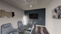 Located at 119 N Pacific Ave in Glendale, California, this single-floor meeting room offers a polished and professional environment designed for focused gatherings and collaborative sessions. The room is defined by a crisp, neutral color palette of soft whites and grays, punctuated by a bold slate-blue accent wall that serves as the visual anchor of the space. Mounted on this feature wall is a large flat-screen display, making the room well-suited for presentations and media-driven meetings. Seating consists of a set of plush, upholstered chairs in a muted blue-gray tone, arranged around sleek glass-top tables with chrome frames, lending the space a contemporary and refined aesthetic. The flooring features patterned carpet tiles in a multi-tone gray scheme, adding visual texture underfoot. Decorative touches include abstract wall art panels and a tall indoor ficus tree, which introduces a natural, organic element to the otherwise structured interior. Recessed ceiling lighting provides even, ambient illumination throughout the room, while a ceiling fan ensures comfortable air circulation. The space is accessed through a standard interior door and benefits from a clean, uncluttered layout that maximizes usability. Overall, the room strikes a balance between functional design and modern sophistication.