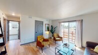 This thoughtfully designed single-floor unit blends mid-century modern aesthetics with contemporary updates throughout. The living room serves as the heart of the space, featuring light hardwood flooring, a textured ceiling, and a large sliding glass door framed by sheer curtains that floods the room with natural light and provides access to an outdoor area. The open layout easily accommodates seating and a glass-top coffee table, creating an inviting and functional gathering space.

Adjacent to the living area is a compact dining nook with hardwood flooring, offering an intimate setting for meals. The kitchen is well-appointed with gray and blue shaker-style cabinetry, gold hardware accents, granite-look countertops, and a full suite of stainless steel appliances including a range, microwave, and refrigerator, all tied together with warm wood flooring.

The unit includes two bedrooms, both carpeted and brightened by windows with outdoor views. One bedroom features a sliding closet door for convenient storage. An additional flexible room with carpet and large windows can serve as a home office or den. The bathroom is finished with wood-look flooring, a tiled shower-tub combo, and matte black fixtures, completing this well-rounded and move-in ready space.