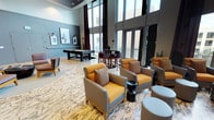 This contemporary amenity lounge is a well-appointed social space designed for both relaxation and recreation. The room immediately impresses with its soaring double-height ceilings and an expansive wall of floor-to-ceiling windows, flooding the interior with natural light and creating an open, airy atmosphere. The soft gray walls and patterned area rug provide a refined, neutral backdrop that complements the bold design accents throughout.

The seating area is thoughtfully arranged with plush armchairs upholstered in warm amber and soft gray tones, accented with textured throw pillows. Small accent tables are positioned throughout for convenience, encouraging conversation and casual gatherings. The layout is open and inviting, offering a natural flow between different activity zones.

Toward the rear of the space, a full-size billiards table anchors the recreation area, accompanied by high-top bar seating with dark wood stools — ideal for socializing and leisure. Framed artwork on the walls adds a curated, gallery-like quality to the environment. Dramatic floor-length curtains in deep burgundy provide a rich contrast to the otherwise cool-toned palette. Together, these elements create a sophisticated yet approachable communal space that balances style with functionality.