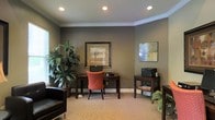 This single-floor office or business center space offers a polished, professional environment designed for productivity and comfort. The room is finished with rich, dark gray walls that create a focused and refined atmosphere, complemented by crisp white crown molding that adds an elegant architectural detail along the ceiling perimeter. Recessed lighting illuminates the space evenly, enhancing the warm, neutral-toned carpeting underfoot. At the center of the room, a dark wood writing desk paired with a cushioned red chair serves as the primary workstation, while a secondary dark wood side table accommodates a printer, making the space fully functional for everyday office tasks. A black leather accent chair is positioned nearby, offering additional seating. Lush indoor plants, including a tall floor plant and a smaller fern, bring a touch of nature into the room, softening the professional aesthetic. Framed artwork adorns the walls, contributing to the curated, welcoming feel of the space. A window allows natural light to filter in, further brightening the interior. Overall, the space strikes a balance between practicality and comfort, making it an inviting and well-equipped environment for work.