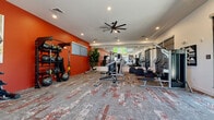 This fitness center offers a comprehensive workout environment spread across a single level, thoughtfully designed to accommodate a wide range of exercise needs. The space is defined by a striking bold orange accent wall that energizes the interior and creates a visually dynamic backdrop for the strength and functional training zones. Patterned carpet in muted tones of grey and red runs throughout, adding texture and visual interest underfoot.

The strength training area is well-stocked with a multi-rack dumbbell station, kettlebells, medicine balls, resistance bands, and TRX suspension trainers, along with a cable machine and adjustable bench. A dedicated cardio section houses multiple Life Fitness treadmills and ellipticals, positioned near bold black-and-white graphic artwork. A large wall-mounted television is centrally positioned for viewing during workouts.

A floor-to-ceiling mirror expands the sense of space and supports proper form during training sessions. A large-bladed ceiling fan ensures comfortable air circulation throughout. Tall glass doors along one wall open to an outdoor terrace with views of surrounding greenery, flooding the space with natural light and providing a seamless connection to the exterior. Potted tropical plants contribute a fresh, lively atmosphere throughout.