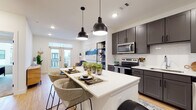 Located in Austin's dynamic East Side neighborhood, this single-story residence at 1503 Robert Weaver Ave offers a thoughtfully designed open-concept layout with contemporary finishes and a clean, modern aesthetic. The kitchen is a true centerpiece, featuring dark gray shaker-style cabinetry, white quartz countertops, a full suite of stainless steel appliances including a French door refrigerator, electric range, built-in microwave, and dishwasher, as well as a white subway tile backsplash. A large central island with seating provides an ideal space for casual dining and entertaining, illuminated by a pair of striking black pendant lights. Warm wood-look flooring flows throughout the main living areas, creating a seamless and inviting atmosphere. The living room is bright and airy, benefiting from natural light streaming through French doors that open to an outdoor balcony, complemented by clerestory windows above. The bedrooms offer plush carpeting, large windows with neighborhood views, and ceiling fans for added comfort. The bathroom is well-appointed with a glass-enclosed shower, wood-look flooring, and dark vanity cabinetry that echoes the kitchen's palette. A built-in mudroom area near the entry adds practical functionality to this well-rounded, move-in-ready Austin residence.