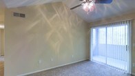 This single-floor space showcases a thoughtfully designed interior that balances comfort and functionality. The standout architectural feature is the vaulted ceiling, which lends a sense of openness and volume to the main living area. A ceiling fan with a light fixture is centrally mounted, providing both air circulation and overhead illumination.

The room is finished with wall-to-wall carpeting in a neutral tone, contributing to a warm and cozy atmosphere underfoot. The walls are painted in a soft, muted yellow-beige hue that reflects light and creates an inviting ambiance throughout the space.

A large sliding glass door fitted with vertical blinds allows abundant natural light to pour in, while also offering a view and potential access to an outdoor area. The interplay of sunlight through the blinds casts interesting patterns across the walls, adding a dynamic visual element to the room.

The space includes multiple electrical outlets along the baseboards, indicating versatility for various furniture arrangements and uses. The clean, simple lines of the interior design provide a neutral canvas that can be easily personalized. Overall, the combination of vaulted ceilings, natural light, and soft finishes creates a bright, airy, and welcoming environment.
