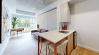 Located in the heart of Montreal at 1201 Rue de la Montagne, this thoughtfully designed studio unit makes excellent use of its compact footprint across a single floor. The open-plan living area is anchored by light hardwood flooring and striking exposed concrete ceilings, lending the space an industrial-modern character. A kitchen island with a white quartz countertop and integrated sink doubles as a breakfast bar with seating for two, seamlessly connecting the kitchen to the living area. White cabinetry, a stainless steel refrigerator, and warm wood accents complete the kitchen. The living area is bright and airy, benefiting from large windows that frame views of lush greenery, allowing natural light to fill the space. A built-in Murphy bed integrated into the cabinetry wall offers a smart space-saving sleeping solution. The hallway connects to a clean, all-white bathroom featuring a vessel sink vanity with ample built-in shelving, a glass-enclosed shower, and light tile flooring. A dedicated laundry area houses a stacked washer-dryer unit, adding everyday practicality. The overall ambiance is modern and minimalist, with carefully chosen details that maximize both functionality and style.