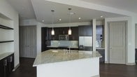 This single-floor space showcases a thoughtfully designed open-concept layout with a strong emphasis on modern aesthetics. The kitchen serves as the centerpiece, featuring rich dark cabinetry that contrasts elegantly against a soft grey subway tile backsplash. A generously sized island with a striking granite countertop in cool white and grey tones provides both a functional workspace and a natural gathering point, complete with an integrated sink and modern faucet. Two pendant lights hang above the island, casting warm, focused illumination that complements the recessed ceiling lighting throughout. Stainless steel appliances, including a French door refrigerator, built-in microwave, and gas range, lend a professional-grade quality to the culinary space. A dedicated wine storage nook with a stone tile accent wall adds a refined touch to one corner. Dark hardwood flooring grounds the space and flows seamlessly throughout, creating a cohesive and polished look. The open design connects the kitchen to the surrounding living areas, enhancing the sense of spaciousness and encouraging an effortless flow between spaces. The overall ambiance is contemporary, clean, and sophisticated.