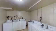 This single-floor laundry room is a practical, utilitarian space designed to accommodate multiple users simultaneously. The room features several Whirlpool top-loading washing machines and matching dryers arranged along the walls, providing substantial laundry capacity. The interior is finished with light-colored wall panels and a smooth ceiling with overhead fluorescent lighting, ensuring the space is well-lit. Exposed plumbing and utility connections are visible along the back wall, reflecting the functional, no-frills nature of the room. The floor is finished with a light-toned linoleum surface, which is easy to clean and maintain. The layout allows for comfortable movement between machines, making the laundry process efficient. Overall, the space is straightforward and purpose-built, prioritizing practicality and accessibility over aesthetics.