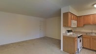 This single-floor space offers a straightforward, functional layout with an open living area that flows naturally toward the entryway. The living area is finished with wall-to-wall neutral carpeting, contributing to a quiet and comfortable atmosphere. Walls are painted in a soft, light tone, keeping the interior feeling clean and understated.

Adjacent to the living area, the kitchen is partially separated by a half-wall partition, providing a degree of definition between the two spaces without fully closing them off. The kitchen features warm medium-toned wood cabinetry on the upper level, offering ample storage. Countertops display a granite-style surface, adding a practical and slightly refined touch to the cooking area. Standard appliances include a gas range and a built-in microwave, both in white finishes that coordinate with the overall neutral palette.

The entryway door is visible from the main living area, underscoring the open sightlines throughout the space. The single-floor layout ensures ease of movement, making this a practical and accessible unit suited for everyday living.