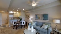 This single-floor residence offers a thoughtfully designed open-concept layout that seamlessly connects the living, dining, and kitchen areas into one cohesive and functional living space. The living area serves as the centerpiece, featuring a comfortable blue-grey upholstered sofa accented with patterned throw pillows in soft greens and whites. A round glass-top coffee table anchors the seating area, while a pair of framed wall art pieces in cool blue tones add a refined decorative touch. A ceiling fan with an integrated light fixture provides both comfort and ambient illumination, complemented by recessed lighting throughout. Adjacent to the living area, the dining space accommodates a dark wood dining table paired with white upholstered chairs, creating an elegant contrast. A fresh floral arrangement adds a welcoming, natural element to the dining setting. The kitchen, visible in the background, features light wood cabinetry, a tile backsplash, and ample counter space, offering a clean and functional cooking environment. Neutral warm-toned walls and light-toned flooring unify the entire space, contributing to an airy, inviting ambiance. The overall aesthetic is clean, modern, and comfortable — ideal for everyday living and casual entertaining.