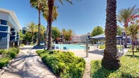 Located at 6730 4th Ave in Sacramento, CA, this resort-style outdoor pool area offers a well-appointed recreational retreat designed for relaxation and social gatherings. The centerpiece is a generously sized, freeform swimming pool with clear turquoise water, complemented by a separate circular spa for additional leisure. The surrounding pool deck is finished in smooth concrete and outfitted with numerous blue lounge chairs and seating arrangements, providing ample space for sunbathing under Sacramento's sunny skies. A covered patio structure offers shaded seating for those seeking relief from the sun, while additional outdoor furniture — including Adirondack chairs and bistro-style tables — is scattered throughout the space for casual dining and socializing. Lush landscaping, including tall palm trees, flowering bougainvillea, and mature shade trees, creates a vibrant, tropical atmosphere that enhances the overall ambiance. A volleyball court and barbecue grilling stations further expand the recreational offerings of this outdoor space. The entire area is enclosed by a wrought-iron perimeter fence, ensuring privacy and security. Residential buildings are visible in the background, situating this pool area as a shared community amenity within a larger residential setting.