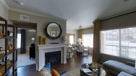 This thoughtfully designed single-floor residence offers an inviting open-concept layout that seamlessly connects the living and dining areas. At the heart of the living room stands a classic white-mantled fireplace, adorned with curated décor including sculptural gold vases and a striking round black-framed mirror — serving as a compelling focal point for the space. Rich, dark hardwood flooring runs throughout, grounding the interior with warmth and continuity. The living area is furnished with a textured sofa accented by vibrant throw pillows, paired with a sleek glass-top coffee table, creating a comfortable yet refined atmosphere. Crown molding along the ceiling adds an elegant architectural touch, while the neutral warm-grey wall palette provides a versatile backdrop for the home's eclectic styling. Adjacent to the living space, the dining area is bathed in natural light streaming through multiple large windows dressed in gold drape panels and horizontal blinds. A round dining table surrounded by upholstered chairs offers an intimate setting for meals. Track lighting overhead provides focused illumination, complementing the natural light. The overall ambiance is sophisticated and welcoming, blending classic architectural details with contemporary furnishings.