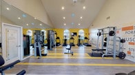 This fitness center offers a thoughtfully designed workout space that combines functionality with an energizing aesthetic. The room is anchored by striking yellow accent walls and a motivational typography mural, creating a vibrant atmosphere conducive to exercise. Soaring vaulted ceilings lend a sense of openness and airiness, amplifying the spacious feel of the facility.

Natural light streams in through arched windows along the far wall, providing views of the outdoors and keeping the space bright and welcoming. Recessed lighting throughout ensures the room is well-illuminated at any time of day. A large wall-to-wall mirror along one side of the room enhances the sense of space while allowing users to monitor their form during workouts.

The flooring features a durable combination of hardwood-style planks accented with bold yellow striping, reinforcing the energetic design theme. The facility is outfitted with a comprehensive range of equipment, including multiple treadmills, elliptical machines, cable weight stations, and free weight racks, catering to a full spectrum of fitness needs. A wall-mounted television provides entertainment during workouts, completing this well-appointed fitness space.
