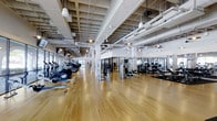 This fitness center offers a well-appointed, single-floor facility designed to accommodate a comprehensive workout experience. The space is defined by its expansive open floor plan, featuring warm hardwood flooring that runs throughout, lending the area a clean and polished aesthetic. Soaring ceilings with an exposed grid structure give the space an industrial yet refined character, while pendant lighting and recessed fixtures ensure consistent, well-distributed illumination across the entire floor.

Along one side of the facility, a row of cardio equipment—including ellipticals, treadmills, and stationary bikes—lines the perimeter, offering users access to natural light streaming in through large windows. The central and opposing areas of the floor are dedicated to strength training, with a selection of cable machines, weight benches, and resistance equipment arranged to allow comfortable circulation between stations.

Floor-to-ceiling mirrors along portions of the walls enhance the sense of openness while providing functional visual feedback during workouts. The overall ambiance is bright, airy, and professionally organized, creating an inviting environment suited for a wide range of fitness activities.