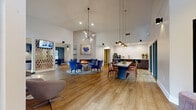 Located at 6316 Quail Hollow Rd in Charlotte, NC, this single-story community clubhouse is a well-appointed gathering space defined by striking vaulted ceilings that lend a sense of openness throughout. The interior is finished with warm wood-look flooring, crisp white walls, and carefully curated contemporary furnishings that create a polished yet welcoming atmosphere.

The main lounge area anchors the space with a sleek linear fireplace set against a full-height stone accent wall, flanked by plush blue velvet accent chairs and a metallic cocktail table atop a patterned area rug. Artwork adds a refined decorative touch above the fireplace surround. An adjoining dining area features a large communal table with pendant lighting overhead and floor-to-ceiling sliding glass doors that open onto a pool courtyard, flooding the space with natural light and providing direct outdoor views.

A dedicated game and recreation zone includes a shuffleboard table, wall-mounted television, and casual seating, along with a compact built-in workstation for quiet tasks. A beverage and refreshment bar with dark cabinetry and countertop service amenities lines one wall near the dining area. Large picture windows throughout frame lush greenery, further connecting the interior to its landscaped surroundings.