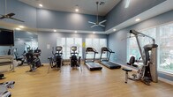 Located at 2250 Sara Jane Pkwy in Grand Prairie, Texas, this single-floor fitness center is thoughtfully designed to support a comprehensive workout experience. The space is finished with warm light hardwood flooring throughout and features calming blue-toned walls that lend a clean, modern aesthetic. Elevated tray ceilings add a sense of spaciousness, while multiple ceiling fans and recessed lighting ensure the environment remains comfortable and well-lit during any workout.

Natural light pours in through several large windows fitted with white horizontal blinds, offering views of the surrounding outdoor area. A full-length mirror along one wall visually expands the space and supports form monitoring during exercise.

The cardio zone is well-equipped with multiple treadmills, elliptical trainers, and stationary bikes, all arranged to maximize usable floor space. The strength training area features a multi-station cable machine, weight benches, and a selection of additional resistance equipment, providing options for both beginners and experienced athletes. A wall-mounted flat-screen television adds entertainment value to the space.

A sanitizing station and clearly marked exit signage reflect attention to health and safety standards. An adjoining room, visible through glass-paneled doors, further extends the facility's functionality. Overall, this fitness center delivers a well-rounded, accessible, and inviting environment for daily exercise.