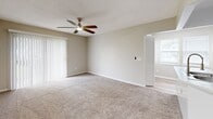 This single-story unit is designed with a clean, neutral aesthetic throughout. The kitchen serves as a central highlight, outfitted with white shaker-style cabinetry extending to the upper walls for ample storage, light-toned countertops, and a full suite of stainless steel appliances including a refrigerator, electric range, and microwave. A kitchen island provides additional counter space and houses a double-basin sink with a flexible faucet, offering both functionality and a natural transition into the adjacent living area.

The living area features plush carpeting, a ceiling fan, and a sliding glass door fitted with vertical blinds that allows natural light to fill the room while providing access to an outdoor space. A secondary room adjacent to the living area also features a sliding glass door, extending the connection to the outdoors.

The bedroom is carpeted and spacious, featuring a walk-in closet visible through a six-panel door. The bathroom includes a white vanity with under-sink storage, a large mirror, pendant lighting, and a separate toilet and tub/shower area with light wood-tone flooring. The overall palette of warm neutrals and white trim creates a cohesive, move-in-ready environment.