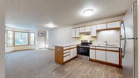 Located at 9950 Redwood St NW in Coon Rapids, Minnesota, this two-bedroom, one-bathroom unit offers a practical and well-organized single-floor layout. The open-concept kitchen and living area forms the heart of the space, where white cabinetry trimmed in warm wood tones pairs with stainless steel appliances — including a range, dishwasher, and refrigerator — and a peninsula with a light countertop that provides additional prep space and casual seating. Light wood-style flooring runs through the kitchen, transitioning to plush carpet in the living area and bedrooms. The living room is highlighted by a curved bay window that invites natural light and offers views of the surrounding greenery, complemented by a wall-mounted air conditioning unit and baseboard heating. Both bedrooms are carpeted and feature ceiling fans, with the primary bedroom offering a generous footprint and direct access to a closet with built-in shelving and a hanging rod. The full bathroom is finished with wood-accented cabinetry, a vanity with a mirror, and a tub-shower combo. Warm wood-toned trim and doors are consistent throughout, lending the space a cohesive and comfortable feel.