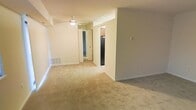 This single-floor residential space offers a clean, functional layout designed for comfortable everyday living. The main living area is finished with wall-to-wall neutral-toned carpeting that lends warmth and continuity throughout the space. A ceiling fan with integrated lighting is centrally positioned overhead, providing both air circulation and ambient illumination to the room.

The open layout connects the living area to adjacent spaces with ease. A doorway leads into the kitchen, which is visible from the main room and features a black refrigerator alongside upper cabinetry, hinting at a practical and well-organized cooking space. A mirrored closet is also visible, offering generous built-in storage that contributes to the overall functionality of the unit.

The walls are finished in a soft, neutral off-white tone that enhances the sense of brightness and openness. Floor-level ventilation registers are integrated into the baseboards, suggesting a central HVAC system for year-round climate comfort. Clean white baseboards frame the carpeted floors throughout, adding a crisp, finished look to the interior. The overall ambiance is calm, understated, and move-in ready, making this space well-suited for those seeking a straightforward and practical living environment.