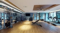 This fitness center in Memphis, TN offers a thoughtfully designed workout environment that balances functionality with contemporary aesthetics. The space features warm hardwood-style flooring throughout, complemented by a combination of crisp white and deep teal-blue accent walls that create an energizing yet refined atmosphere. Recessed lighting and LED strip accents provide consistent, well-distributed illumination across the floor.

The strength training area is comprehensively outfitted with a power rack loaded with olympic weight plates, a multi-functional cable machine, a GymRAX functional training rig complete with battle ropes and suspension equipment, and a tiered dumbbell rack offering a full range of free weights. An adjustable weight bench provides versatility for various exercises.

The cardio section is positioned along a wall of large windows that invite abundant natural light and outdoor views of the surrounding Memphis streetscape. Multiple treadmills, stationary bikes, and an elliptical trainer are available for aerobic training. A wall-mounted flat-screen television adds entertainment options.

Architectural details include a distinctive wood-paneled ceiling inset with a large ceiling fan, exposed industrial-style ductwork, and floor-to-ceiling glass entry doors. Built-in shelving provides practical storage within the space.