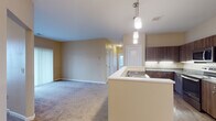 This single-floor residence is designed with an open, functional layout that connects the kitchen and living area seamlessly. The kitchen is well-appointed with dark wood cabinetry, granite countertops, and a full suite of stainless steel appliances, including a refrigerator, range, and over-the-range microwave. Light wood-look flooring runs through the kitchen area, while pendant lighting above the peninsula adds a refined touch. The peninsula itself features a built-in sink, offering a practical workspace that overlooks the living area.

The living space is generously sized, finished with neutral-toned carpet that extends throughout the bedrooms. Each bedroom is bathed in natural light through blinds-fitted windows, providing views of the surrounding neighborhood. The rooms feature warm beige walls and clean white trim, creating a calm and neutral aesthetic.

The bathroom includes a dark wood vanity with a granite countertop, light wood-look flooring, and white panel doors, maintaining a consistent design language found throughout the unit. Overall, the space presents a cohesive, neutral palette with practical finishes, offering a comfortable and functional living environment across all areas.