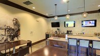 This single-floor hospitality space is designed to offer guests a comfortable and functional environment for dining and relaxation. The room centers around a wooden island equipped with a sink, counter seating, and ample workspace, making it ideal for serving and self-service dining. Dark wood dining tables and upholstered chairs are arranged throughout the space, providing seating for multiple guests simultaneously.

The walls are adorned with a textured geometric wallcovering that adds visual interest and a contemporary touch to the room. Crown molding with a dark wood border runs along the upper walls, lending a refined, structured look to the interior. Two globe pendant lights hang above the island, providing focused illumination and a decorative accent that complements the overall design.

A beverage station along the back wall includes coffee dispensers and accompanying service items, catering to guests' refreshment needs. Two wall-mounted flat-screen televisions are positioned at viewing angles accessible from multiple seating areas, enhancing the guest experience. Recessed ceiling lighting throughout ensures the space is well-lit, while neutral tones on the walls and tile flooring contribute to a clean, welcoming ambiance suited to a hotel or hospitality setting.