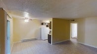 This single-floor residence offers a straightforward and functional layout designed for everyday living. The main living area is open and spacious, featuring light-toned tile flooring that extends throughout the space, a ceiling fan for air circulation, and a baseboard heating unit along the wall. A textured popcorn ceiling adds a classic touch overhead. The kitchen is partially separated from the living area by a half-wall partition, creating a semi-open feel while maintaining distinct zones. It is equipped with dark wood cabinetry, a white refrigerator, and a countertop workspace, providing practical storage and meal preparation space. The warm mustard-yellow accent wall adds a pop of color and helps define the boundary between the kitchen and living spaces. A doorway visible toward the back of the unit leads to the remaining rooms, suggesting additional private living quarters such as a bedroom or bathroom. The overall ambiance is utilitarian and functional, with a neutral color palette that offers a blank canvas for personalization. The space is well-suited for those seeking a manageable, easy-to-maintain single-level home.