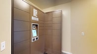 This space features a Package Concierge automated locker system, providing a secure and convenient solution for package delivery and retrieval. The locker bank, finished in a warm bronze-tone panel design, spans floor to near-ceiling height and wraps around a corner, maximizing storage capacity within the compact alcove. The unit is equipped with an integrated touchscreen interface and barcode scanner, allowing recipients to retrieve their deliveries independently at any hour. The surrounding walls are painted in a neutral cream tone with clean white baseboards, contributing to a tidy, well-maintained environment. The system's smart design eliminates missed deliveries and ensures packages remain secure until picked up, making it a practical and modern amenity for any multi-resident or commercial building.