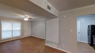 This single-floor residence presents a clean, functional layout finished with warm hardwood flooring that runs throughout the main living area, adding a sense of continuity and warmth. The walls are painted in soft, neutral gray tones that complement the bright white trim, baseboards, and crown molding — classic details that lend the space a polished character.

The main living area is open and airy, anchored by a ceiling fan that provides both light and airflow. A white entry door welcomes you at the front, and the space transitions naturally toward interior rooms via a defined architectural division featuring built-in storage or utility access panels. HVAC vents are strategically placed both in the ceiling and near the floor, ensuring efficient climate control throughout.

The textured ceiling adds a layer of visual depth to the space. The layout appears straightforward and easy to navigate, with clear sightlines from the entry through the main living zone. Overall, the space offers a practical, move-in-ready environment with modest but well-maintained finishes, making it a comfortable and versatile setting suited to a variety of living arrangements.