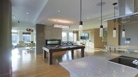 This modern, single-floor space is designed for social gathering and leisure, showcasing an open-concept layout that seamlessly connects multiple functional zones. At the heart of the space sits a sleek billiards table, positioned beneath a striking rectangular pendant light fixture and surrounded by warm hardwood flooring that extends throughout. A textured stone accent wall serves as a dramatic focal point, adding depth and visual interest to the interior.

The lounge area beyond features comfortable seating arranged around a flat-screen television, creating an inviting media zone ideal for relaxation. Natural light streams in through large windows, brightening the space and offering views of the surrounding outdoor environment. A granite-topped counter is prominently positioned in the foreground, suggesting a refreshment or serving area that enhances the space's entertainment functionality.

Contemporary pendant lighting in varied styles — including cylindrical and textured glass designs — hang at different heights, contributing to the layered, curated aesthetic. Recessed ceiling lighting provides consistent ambient illumination throughout. Warm, neutral tones in the walls and furnishings are complemented by dark wood accents, striking a balance between modern sophistication and approachable comfort. The open floor plan ensures excellent sightlines across the entire space.