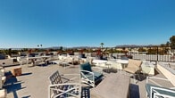 Located in the heart of Los Angeles, this rooftop terrace at 418 N Norton Ave offers an impressive open-air experience defined by sweeping panoramic views of the Hollywood Hills, surrounding palm tree-lined streets, and the expansive LA cityscape. Bathed in abundant Southern California sunshine, the terrace is thoughtfully designed as an outdoor communal retreat with multiple distinct seating zones. White powder-coated lounge chairs, armchairs, and café-style tables are arranged throughout the deck to accommodate both relaxed lounging and casual conversation. Sun loungers provide dedicated areas for soaking in the California rays, while decorative planters featuring vibrant red cordyline and lush greenery add a polished, resort-like ambiance to the space. The clean, contemporary concrete deck surface connects the various seating areas seamlessly, and a sleek black metal perimeter railing ensures unobstructed sightlines across the horizon. The terrace benefits from an open layout that maximizes the sense of space and takes full advantage of the clear blue skies characteristic of the region. Whether used for social gatherings or quiet relaxation, this rooftop destination delivers an exceptional outdoor experience elevated above the Los Angeles urban landscape.