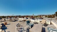 Located in the heart of Los Angeles, this rooftop terrace at 418 N Norton Ave offers an impressive open-air experience defined by sweeping panoramic views of the Hollywood Hills, surrounding palm tree-lined streets, and the expansive LA cityscape. Bathed in abundant Southern California sunshine, the terrace is thoughtfully designed as an outdoor communal retreat with multiple distinct seating zones. White powder-coated lounge chairs, armchairs, and café-style tables are arranged throughout the deck to accommodate both relaxed lounging and casual conversation. Sun loungers provide dedicated areas for soaking in the California rays, while decorative planters featuring vibrant red cordyline and lush greenery add a polished, resort-like ambiance to the space. The clean, contemporary concrete deck surface connects the various seating areas seamlessly, and a sleek black metal perimeter railing ensures unobstructed sightlines across the horizon. The terrace benefits from an open layout that maximizes the sense of space and takes full advantage of the clear blue skies characteristic of the region. Whether used for social gatherings or quiet relaxation, this rooftop destination delivers an exceptional outdoor experience elevated above the Los Angeles urban landscape.