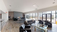 This contemporary clubroom in Houston, TX is thoughtfully designed to offer a dynamic and versatile social environment across a single open floor. The space is anchored by a dramatic floor-to-ceiling stacked stone accent wall housing a sleek linear fireplace, flanked by plush emerald green velvet seating and a rustic natural wood coffee table. Large steel-framed glass doors and expansive windows flood the interior with natural light and open directly onto an outdoor terrace with open city views, seamlessly blending indoor and outdoor living. A dedicated lounge area features leather sofas with gold-accented bases arranged around a wall-mounted television, ideal for gatherings. The game area offers both a regulation pool table beneath a striking multi-arm sculptural chandelier and a full ping-pong table, catering to a range of recreational activities. The well-appointed kitchen includes light wood cabinetry, granite countertops, stainless steel appliances, a large French-door refrigerator, and a generous marble-topped island with modern chrome bar stools. Decorative artwork panels add a refined artistic touch throughout the kitchen area. Wide-plank hardwood flooring runs throughout the main entertainment zones, complemented by large-format tile in the kitchen, tying the entire space together with a cohesive, modern aesthetic.