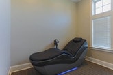 Located at 299 Harris Ln in Gallatin, Tennessee, this single-floor wellness facility offers a dedicated treatment space designed for therapeutic relaxation and recovery sessions. The room centers around a sleek, modern therapy pod finished in black upholstery, complete with an integrated ambient blue LED base and a built-in headrest pillow. A mounted tablet display positioned beside the pod allows clients to engage with guided content or media during their session. The room is finished with neutral warm-toned walls and patterned carpet flooring that contribute to a calm, professional atmosphere. A large window fitted with white horizontal blinds and transom glass above allows natural light to filter softly into the space, creating a bright yet controlled environment. Crown molding and clean white baseboards throughout add a polished architectural detail. A wall-mounted metal rack near the entry provides convenient storage, while the overall layout is thoughtfully arranged to maximize comfort and accessibility. The space is compact yet well-appointed, offering a focused, private environment well-suited for wellness treatments and therapeutic services.
