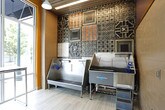 This single-floor pet spa is thoughtfully designed to accommodate both grooming and recreational needs for pets. The space is accessed through a sleek glass-paneled entry door framed in polished metal, flanked by subway-tiled walls in a neutral beige tone that establish a clean, professional first impression.

The grooming area is a standout feature, equipped with stainless steel bathing tubs and a grooming table positioned against a striking decorative wall of mixed metallic and patterned tiles. Large floor-to-ceiling windows flood this area with abundant natural light and provide open views of the surrounding streetscape, creating an airy, welcoming environment. Light wood-toned flooring ties the space together with warmth.

Adjacent to the grooming station is an expansive activity area finished with polished concrete flooring, offering a durable and easy-to-maintain surface suited for active use. Bold mustard-yellow walls are accented with playful illustrated animal silhouettes and descriptive mood words, lending energy and personality to the room. Pendant lights hang from a drop ceiling, and a wall-mounted television adds a functional touch. Red activity equipment is positioned throughout, and large storefront windows along one wall maintain a strong visual connection to the outdoors.