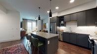 This single-floor residence showcases a thoughtfully designed open-concept layout with contemporary finishes throughout. The kitchen serves as a central highlight, featuring gray shaker-style cabinetry extending to the ceiling, stainless steel appliances, white subway tile backsplash, and a granite-topped island with seating for multiple guests. Pendant lighting with sleek cylindrical fixtures illuminates both the island and surrounding workspace, while warm hardwood flooring flows throughout the main living areas.

The living room is defined by floor-to-ceiling glass doors opening to a private balcony, inviting natural light and broad city views. The sitting area accommodates a rich, tufted leather sofa complemented by geometric-patterned rugs, creating a layered, eclectic ambiance.

A secondary room with neutral carpet, a double-door closet, and wall décor provides flexibility as a bedroom or home office. The primary bedroom offers carpeted flooring and a large window framing expansive outdoor views.

The bathroom features granite-topped gray cabinetry, tile flooring, and clean white walls, while an additional bath window captures sweeping city panoramas, adding a distinctive and luminous quality to the space overall.