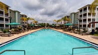 This outdoor amenity area centers around a generously sized resort-style swimming pool with striking turquoise water, creating a vibrant focal point within the community. The pool is elongated in design, offering ample space for both lap swimming and leisurely use. Surrounding the pool, a broad tiled deck provides a clean, durable surface finished in warm earth tones that complement the overall aesthetic.

On both sides of the pool, rows of lounge chairs are neatly arranged, each paired with teal market umbrellas that provide shade throughout the day. Classic black lamp posts are positioned along the perimeter, ensuring the space is well-lit during evening hours. Tall, slender palm trees are planted throughout the deck area, adding a tropical character and contributing to the resort-like ambiance of the space.

The pool area is enclosed by a fence, maintaining privacy and security. Lush greenery and mature trees are visible in the background, softening the transition between the pool area and the surrounding buildings. Multi-story residential buildings with balconies flank the space on either side, framing the pool and contributing to a sense of enclosure. The overall atmosphere is inviting, well-maintained, and designed for outdoor relaxation and recreation.