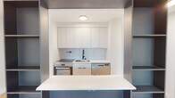 This thoughtfully renovated single-floor unit showcases a clean, modern aesthetic throughout. The open-concept living area is anchored by light hardwood flooring that extends seamlessly across the space, complemented by crisp white walls and recessed lighting that create a bright, airy atmosphere. Large windows frame views of the surrounding urban landscape and treetops, allowing natural light to fill the room while baseboard heating ensures year-round comfort.

The kitchen serves as a striking focal point, framed by a bold dark-toned shelving unit that doubles as a room divider and display feature. Inside, white flat-panel cabinetry reaches to the ceiling, paired with a classic subway tile backsplash and a white countertop that extends into a breakfast bar. Stainless steel appliances include a range with oven and a dishwasher, accompanied by a matte black faucet that adds a contemporary accent.

The bathroom continues the modern design language with light hardwood-style flooring, white subway tile, a wall-mounted vanity with integrated drawers, a mirrored medicine cabinet, and a sleek matte black faucet. The overall design of this space emphasizes minimalist style, functional storage, and a cohesive neutral palette throughout.