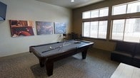This single-floor game room is thoughtfully designed as a social and recreational space. At its center sits a full-size pool table with a slate-gray felt surface and rich dark wood finish, positioned on textured carpeted flooring that adds warmth and comfort underfoot. The room is illuminated by a combination of recessed ceiling lighting and generous natural light streaming in through large windows fitted with horizontal blinds, offering views to an outdoor balcony or walkway beyond. The neutral-toned walls provide a clean backdrop, accented by a striking set of vintage automotive canvas prints that lend the space a casual, retro-inspired character. A small bistro-style table with accompanying chairs is tucked near the window, offering a spot for spectators or casual conversation. The overall layout is open and functional, allowing ample room to move around the pool table with ease. The space strikes a balance between a welcoming recreational environment and a well-appointed amenity area, making it well-suited for socializing and leisure. The cohesive design elements — from the wall art to the furnishings — create an inviting atmosphere that encourages guests or residents to gather and enjoy the space.