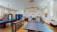 Located at 2201 Greenville Blvd in Greenville, NC, this single-floor community space is thoughtfully designed to accommodate recreation, relaxation, and productivity under one roof. The open-concept layout is anchored by warm hardwood flooring and soaring vaulted ceilings that lend an airy, expansive feel throughout.

The recreation zone is a standout feature, equipped with a regulation ping-pong table and a billiards table positioned beneath stylish pendant lighting. A comfortable lounge area invites gathering, with a large sectional sofa, accent chairs, and a marble-top coffee table arranged around a sizable flat-screen TV mounted on a textured tile accent wall. Decorative shelving and motivational wall art add personality to the space.

Natural light floods the interior through a series of tall, narrow windows positioned high along the walls, offering views of the surrounding greenery while maintaining an uplifting atmosphere. The co-working section features individual study nooks with built-in desks, task lighting, and a stone tile surround, providing a focused environment for work or study. A high-top communal table with bench seating offers an additional collaborative workspace.

Modern linear pendant fixtures and recessed lighting throughout ensure the space is well-lit at all times. The overall ambiance is contemporary, welcoming, and multifunctional — designed to serve a diverse range of activities and users.
