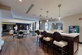 Located in Vienna, Virginia, this sophisticated single-floor clubhouse at Crianza Place offers a well-appointed communal space designed with modern elegance. Rich wood-look flooring flows seamlessly throughout, anchoring a cohesive and refined aesthetic.

The first living area serves as an inviting lounge, featuring plush white seating accented with red geometric throw pillows and a sleek black side table. Floor-to-ceiling windows with dramatic dark curtains flood the space with natural light and open directly to an outdoor patio with seating — ideal for relaxation and social gatherings.

The second living area provides a more intimate setting, outfitted with deep burgundy armchairs and a sofa arranged around a glass-top coffee table. A large flat-screen TV is mounted within a custom dark wood built-in wall unit, while recessed lighting creates a warm, welcoming ambiance.

The dining area features a round wood table surrounded by cream upholstered chairs, complemented by vibrant framed artwork on neutral walls — perfect for private meetings or casual dining.

Additional highlights include a built-in banquette seating nook with bold patterned upholstery, a business center alcove, and a textured sculptural accent wall. The space is accessible directly from the outdoor courtyard, seamlessly blending indoor comfort with outdoor living.