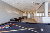The Pointe, located at 1401 College Ave in Manhattan, Kansas, is a well-appointed community amenity space designed for social recreation and leisure. The single-floor layout is open and generously proportioned, offering a welcoming atmosphere for residents to gather and unwind.

The main game room is the centerpiece of the space, finished with neutral gray walls accented by bold lettered signage reading "THE POINTE." The room is outfitted with a full suite of game tables, including a foosball table, a shuffleboard table, and a billiards table, providing a variety of entertainment options. Cornhole boards are also available, adding to the casual, community-oriented feel of the space.

A distinctive white wainscoted divider wall with decorative columns and a dark countertop runs through the interior, creating a subtle separation between areas while maintaining an open sightline throughout. Recessed lighting set into a drop tile ceiling provides consistent, even illumination across the entire space. Durable patterned carpet flooring completes the look, balancing comfort with practicality.

Natural light filters in through multiple double-hung windows along the far wall, brightening the space and offering views to the outdoors. The overall design strikes a balance between polished and approachable, making The Pointe an ideal hub for community interaction at this Manhattan, KS location.