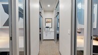 This thoughtfully designed single-floor amenity space combines focused work environments with fitness facilities, catering to a variety of daily needs. At the heart of the workspace area, a central corridor lined with sleek glass-panel sliding doors provides access to a series of private work pods. Each pod is finished with soft blue textured wallcovering, a built-in white desk surface, and an ergonomic rolling chair — creating a quiet, distraction-free setting ideal for individual focus or virtual meetings. Recessed lighting and decorative desk lamps add a warm, welcoming ambiance to each pod. At the end of the corridor, a shared support station features white shaker-style cabinetry, a multifunction printer, and a framed cityscape print that adds a local character to the space. The carpeted flooring throughout the work area features a neutral, multi-tone pattern that complements the contemporary aesthetic. Adjacent to the workspace, a spacious fitness center is visible through large floor-to-ceiling glass windows. The gym is outfitted with a comprehensive selection of cardio and strength-training equipment, including treadmills, ellipticals, and cable machines. Ceiling fans, recessed lighting, and bold navy accent walls create an energizing atmosphere, while the polished concrete floors in the adjoining corridor reflect the natural and artificial light throughout.