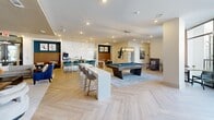 Located at 2221 Berryhill Rd in Charlotte, NC, this thoughtfully designed clubhouse offers a generously sized, single-floor gathering space that blends modern aesthetics with functional amenities. The signature herringbone-patterned hardwood flooring flows throughout, anchoring the space with warmth and visual continuity.

The main living area serves as the centerpiece, featuring a bold teal built-in shelving unit housing a flat-screen TV, flanked by curated décor and surrounded by a mix of plush sofas and accent chairs in a sophisticated neutral palette. Cove lighting with recessed fixtures adds depth and ambiance to the ceiling above.

A dedicated game area includes a full-size billiards table and comfortable lounge seating, while a stylish bar-height communal table with pendant lighting provides additional gathering space alongside a coffee station. Private semi-enclosed dining booths lined with deep navy velvet seating and framed by a striking textured stone accent wall offer intimate nooks ideal for small meetings or casual conversation.

The fitness area is bathed in natural light through floor-to-ceiling windows and is equipped with exercise balls, resistance bands, and a wall-mounted display. A well-proportioned hallway connects the various zones, adorned with contemporary artwork and clean white walls, completing this polished and versatile clubhouse.