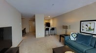 This single-floor unit is thoughtfully arranged to maximize functionality across its open-concept layout. The living area serves as the central hub of the space, featuring neutral-toned carpeting and enough room to comfortably accommodate a seating arrangement. A teal sofa and dark accent furniture add a touch of contrast against the warm beige walls and light-colored textured ceiling, creating an inviting atmosphere.

The kitchen is positioned adjacent to the living and dining areas, separated by a raised breakfast bar that accommodates two bar stools, making it ideal for casual dining or entertaining. The kitchen is equipped with dark appliances, including a refrigerator, and features warm wood-toned cabinetry. A separate dining area with a table and chairs sits nearby, offering a dedicated space for meals.

The bedroom is generously proportioned, providing ample space for a full-sized bed and additional furnishings. The bathroom includes standard fixtures and is conveniently located off the main corridor. The entry area connects all the rooms efficiently, ensuring a logical and practical flow throughout the unit. Overall, the space presents a functional and comfortable living environment with a cohesive, neutral design palette.