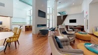 This single-floor amenity lounge is a thoughtfully designed communal space that blends modern aesthetics with comfortable, inviting furnishings. The space is anchored by warm hardwood flooring that extends throughout, complemented by soaring double-height ceilings that lend an airy, expansive feel to the interior.

A striking white brick fireplace column serves as the visual centerpiece of the main lounge area, topped with a wall-mounted television and flanked by an arrangement of tufted gray armchairs and contemporary accent seating in teal and charcoal tones. A bold, oversized yellow sectional sofa lines one wall, adorned with an eclectic mix of decorative pillows in teal, coral, and botanical prints, set against a textured three-dimensional white wall panel that adds depth and visual interest.

Floor-to-ceiling black-framed windows and glass doors bathe the space in natural light while offering views of the surrounding outdoor area, which features palm trees and an exterior lounge. A sculptural indoor planter installation showcases three mature fiddle-leaf fig trees set in walnut-finished planters atop a river rock bed, bringing a natural, organic element indoors.

A sleek white reception or presentation counter anchors the central zone, while a bar-height counter with seating and a geometric diamond-patterned wood accent wall define a distinct social nook. Multiple flat-screen televisions, recessed lighting, and carefully curated décor complete this vibrant, multi-functional gathering space.